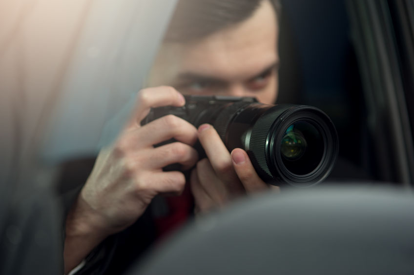 Person with short hair holds a camera, aiming through a car window, appearing to take a photograph discreetly.